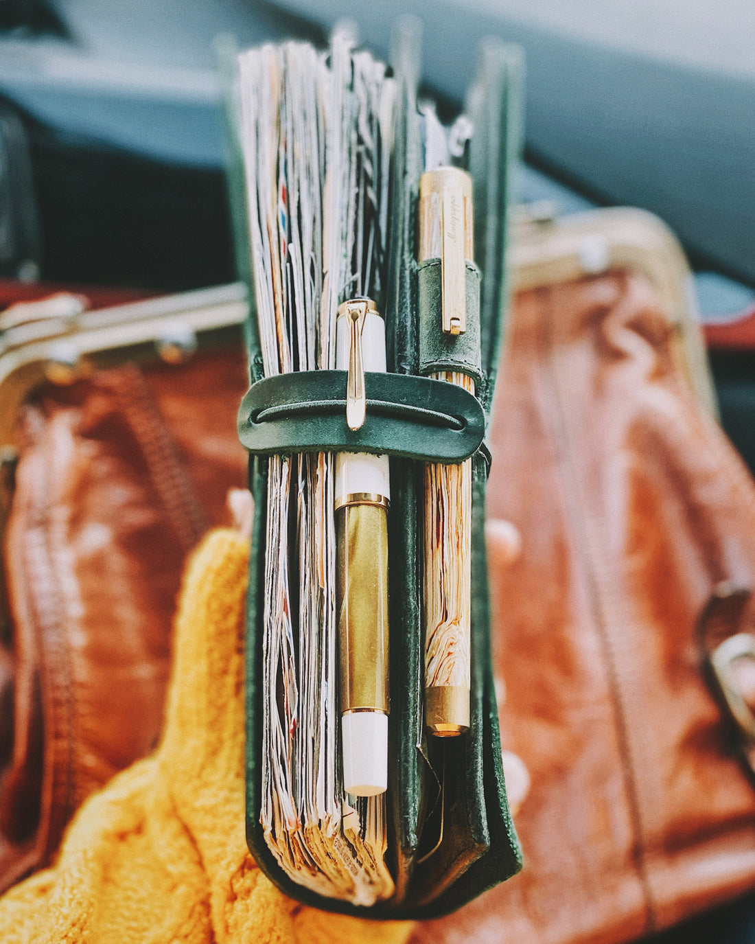 A close-up of a dark green leather journal cover filled with a notebook, a gold fountain pen from Montegrappa and another fountain pen from Pelikan, held in a hand against a blurred background of a brown vintage leather bag and a yellow knit sweater.